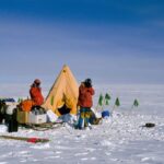 Photo of arctic with three parka clad people and supplies and a yellow tent